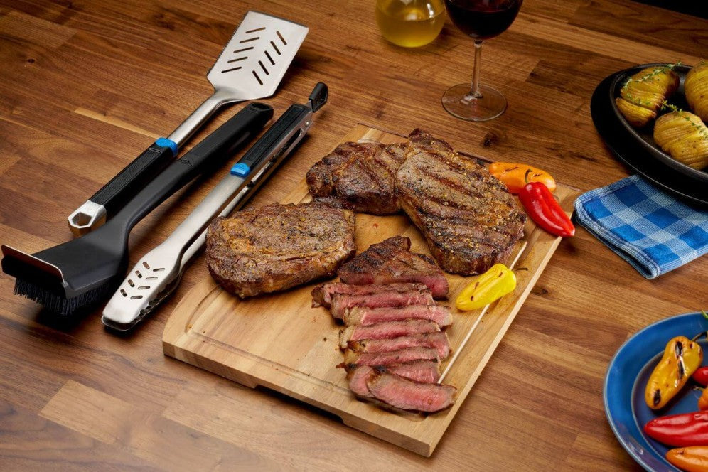 Stainless steel grilling tool set with spatula, tongs, and brush alongside grilled steaks on a wooden cutting board.